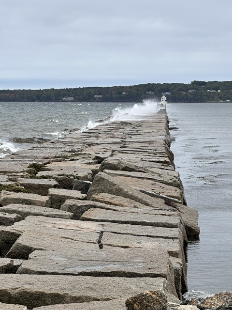 A long granite breakwater extending across the water toward a small lighthouse, with waves breaking against the stones